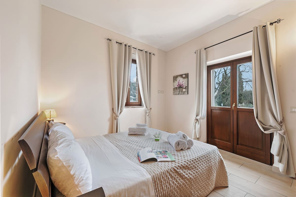 Una cama bien hecha con una revista, toallas y una pequeña planta. Hay una ventana con cortinas, un cuadro en la pared y una puerta con cortina.