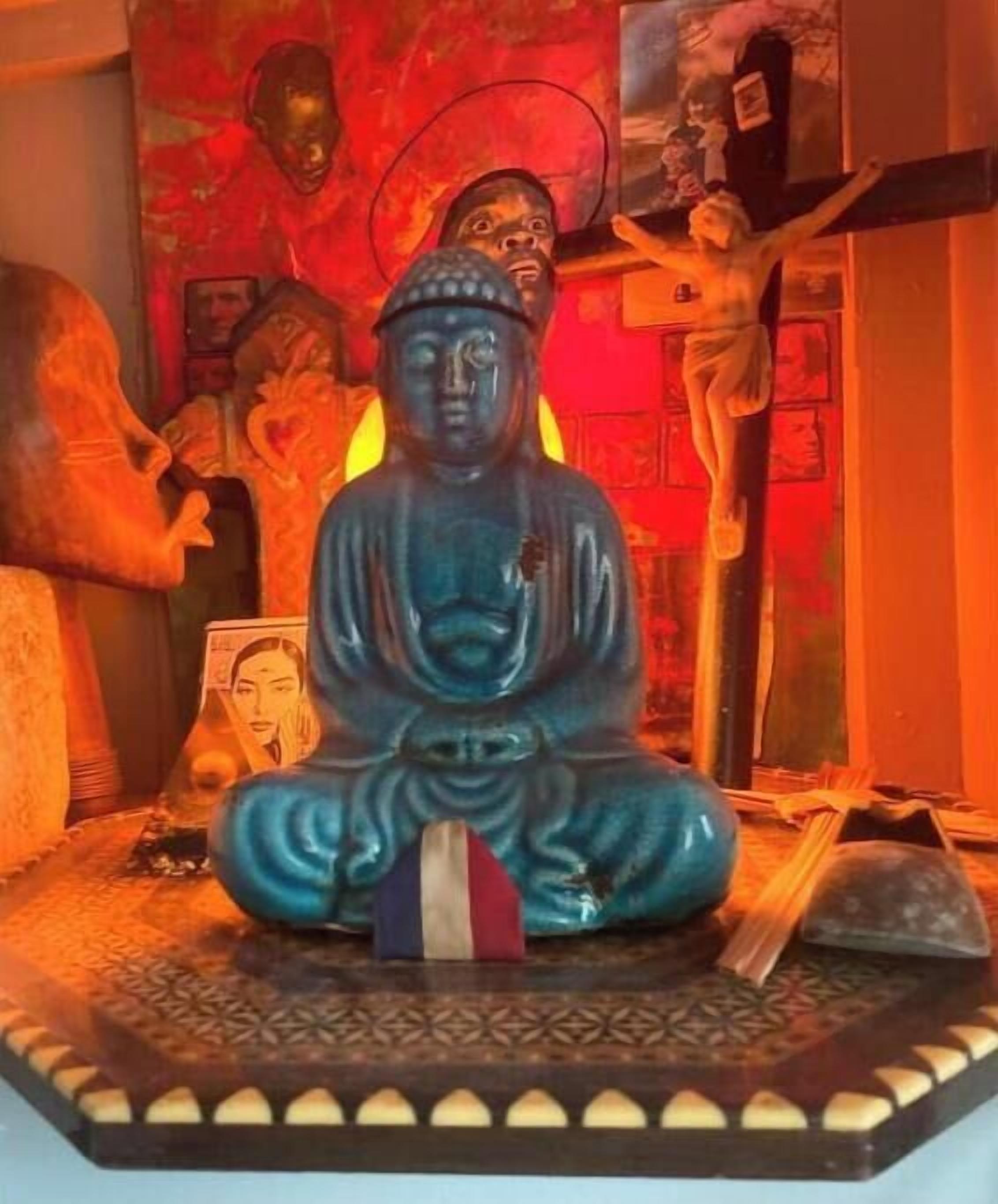 Une statue représentant une figure assise, placée sur un piédestal orné d’une base à motifs, et entourée de divers objets et éléments décoratifs.