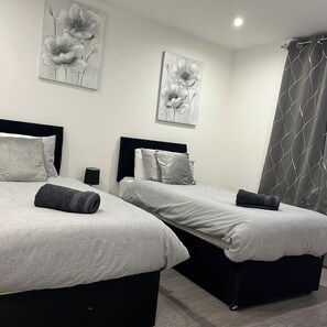 A hotel room with two beds, each with a black headboard, grey bedding, and a rolled towel on the bed. There are two wall-mounted paintings of flowers, and a grey curtain with a wavy pattern. - Free parking Sleek Oasis, 2 Bed, Croydon, Sleeps 6 (Greater London)