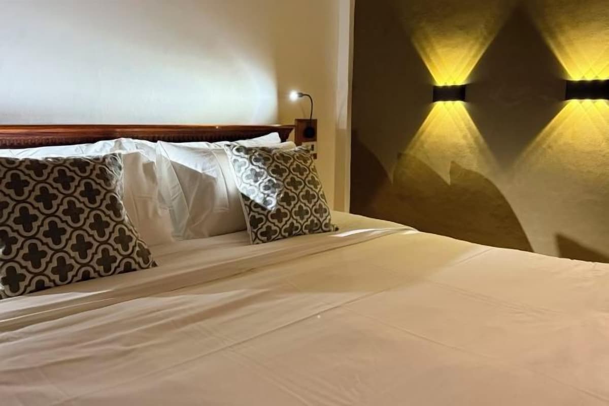 Una cama bien tendida con almohadas estampadas y una lámpara de pared.