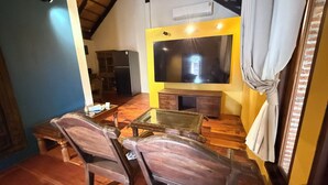 A living room with a wooden floor, a glass coffee table, two wooden chairs, a television, and a fireplace. - Kawarin Kanpuri Villas (Kanchanaburi)
