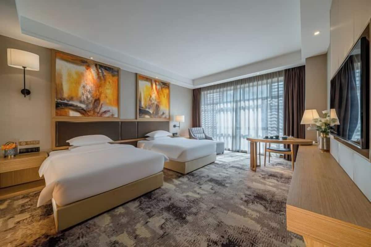 Quarto de hotel com duas camas, uma escrivaninha, televisão e obras de arte nas paredes.