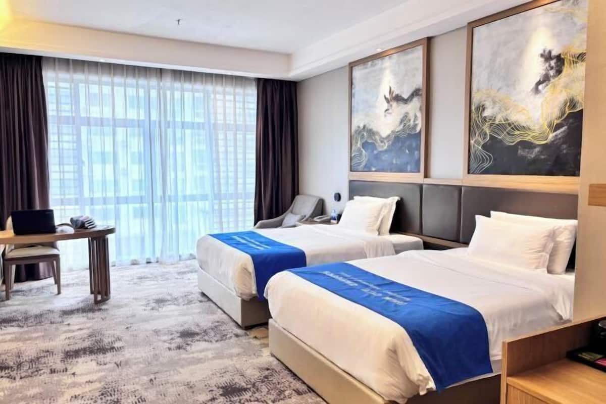 Quarto de hotel com duas camas, uma escrivaninha e uma janela grande com cortinas.