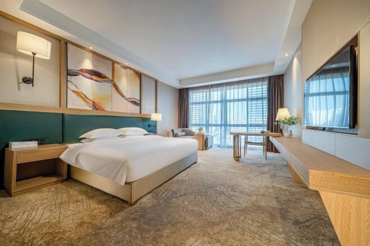 Quarto de hotel com cama, mesas de cabeceira, escrivaninha, cadeira, televisão e uma grande janela com cortinas.