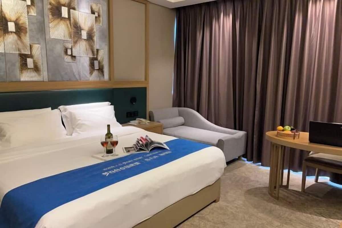 Quarto de hotel com cama, escrivaninha, cadeira, televisão e uma grande janela com cortinas.