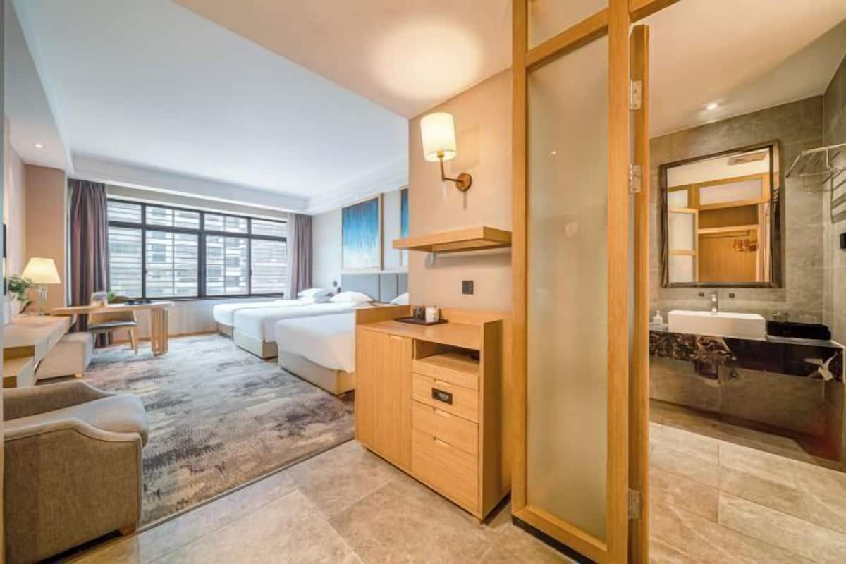 Quarto de hotel com cama, escrivaninha, cadeira, sofá, banheiro com pia e espelho, e uma janela grande.