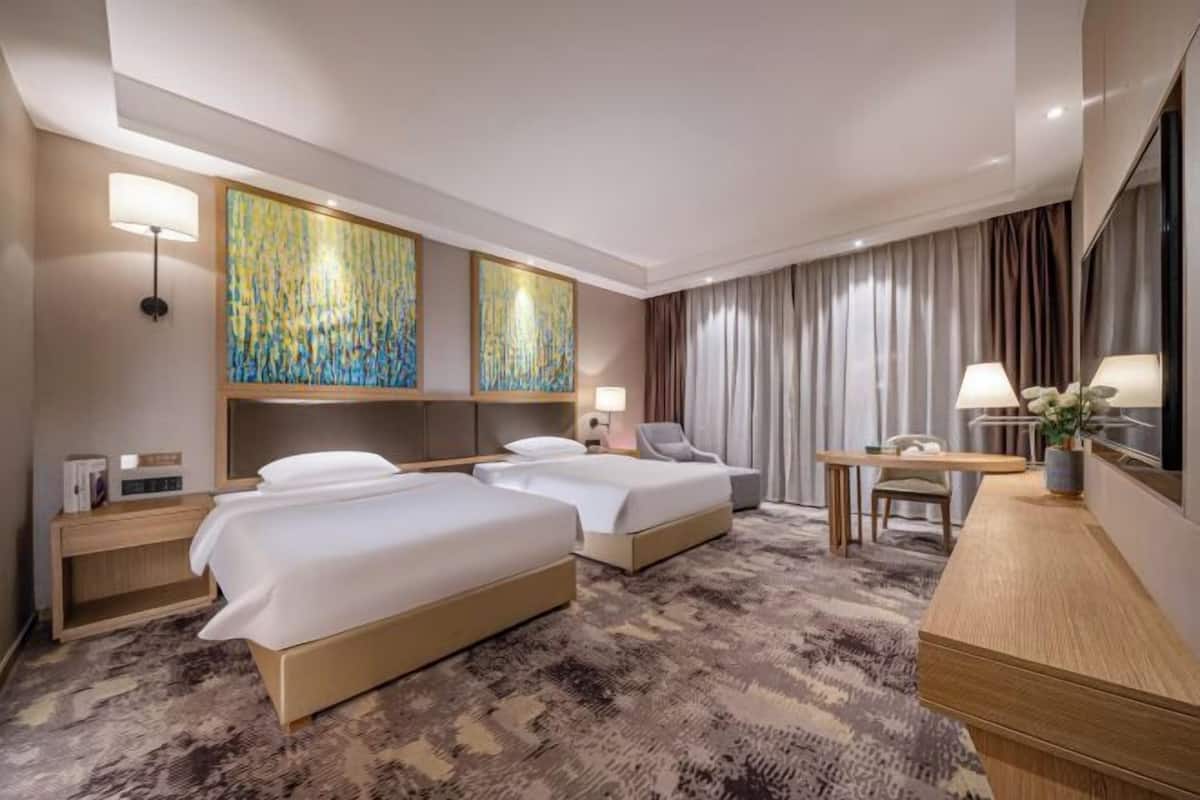 Quarto de hotel com duas camas, uma escrivaninha, uma cadeira, um abajur e obras de arte na parede.