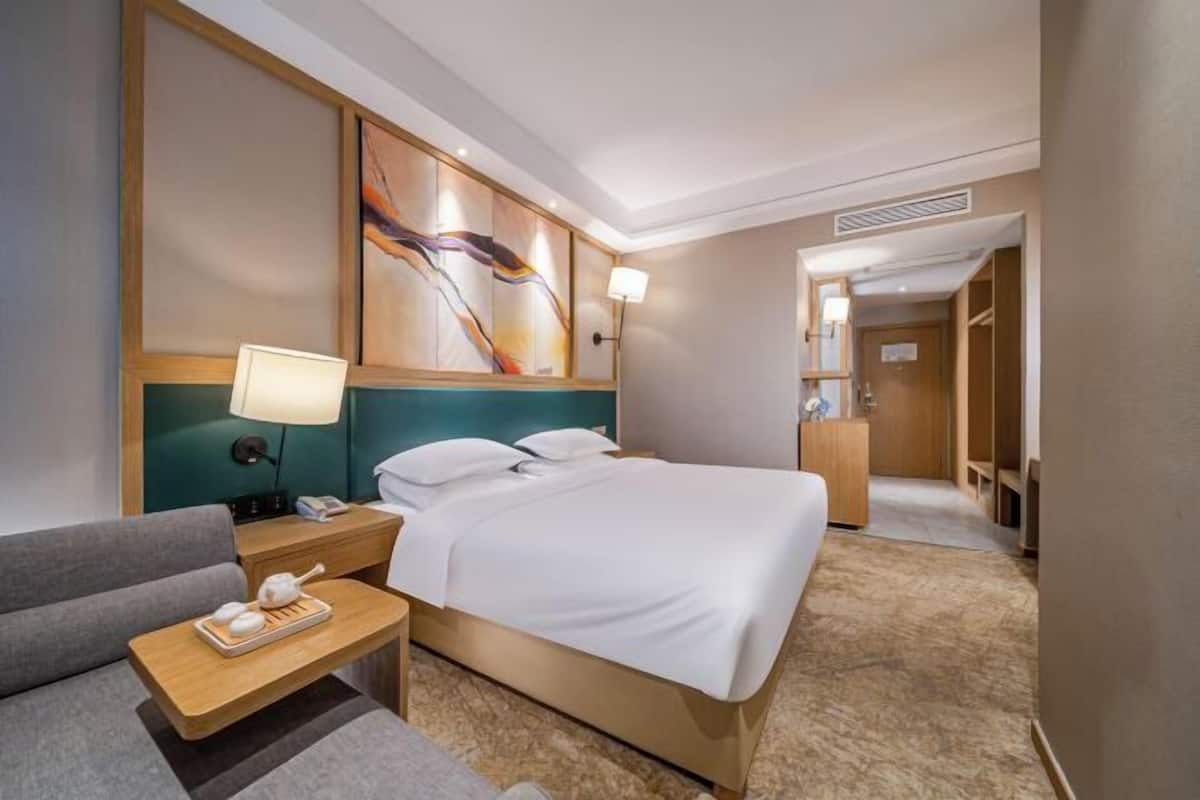 Quarto de hotel com uma cama, uma mesa de cabeceira com abajur, uma cadeira, um quadro na parede e um guarda-roupa.