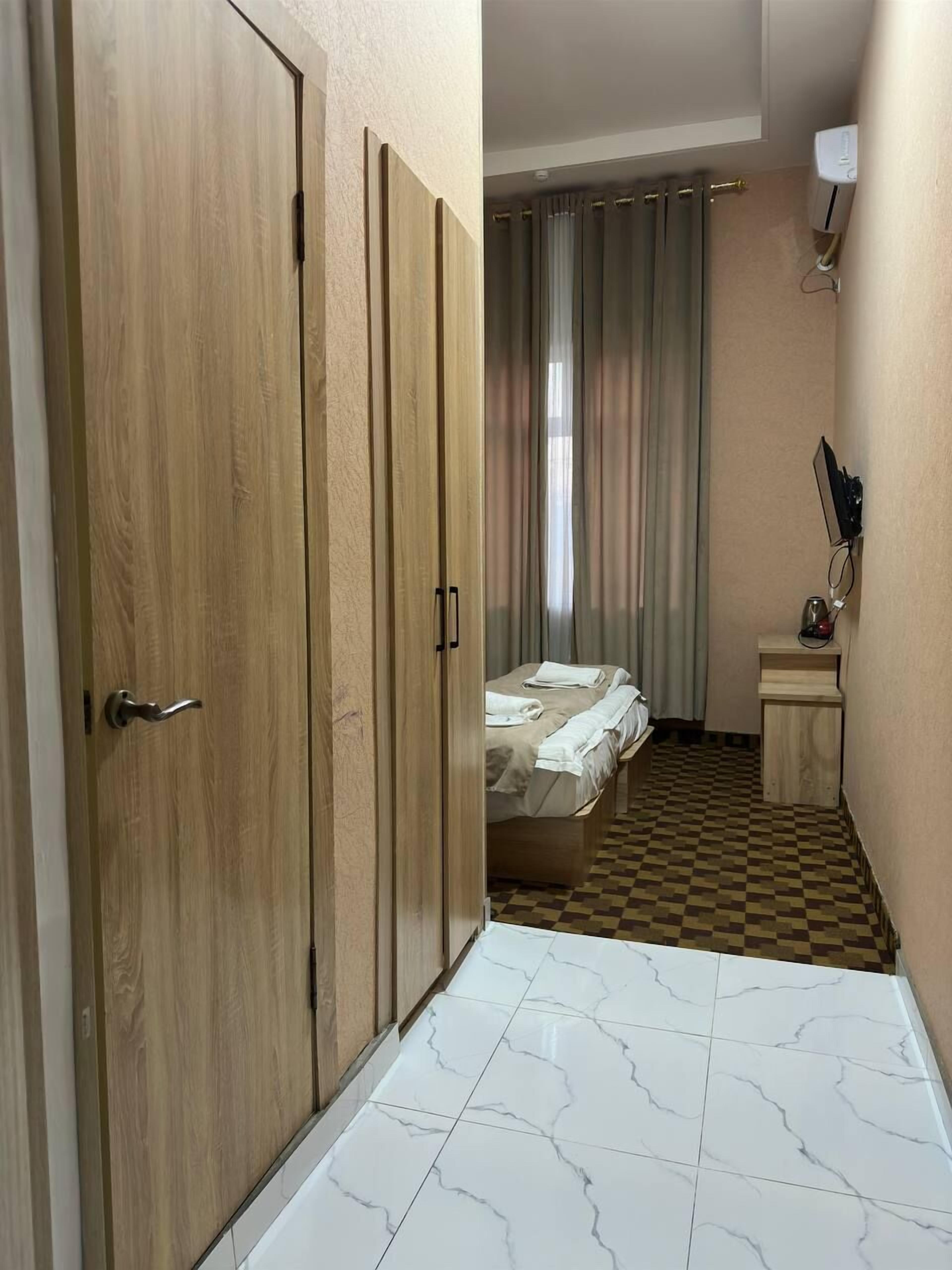 A hotel room with a wooden wardrobe, a bed with a checkered floor, a small bedside table, and a window with curtains.