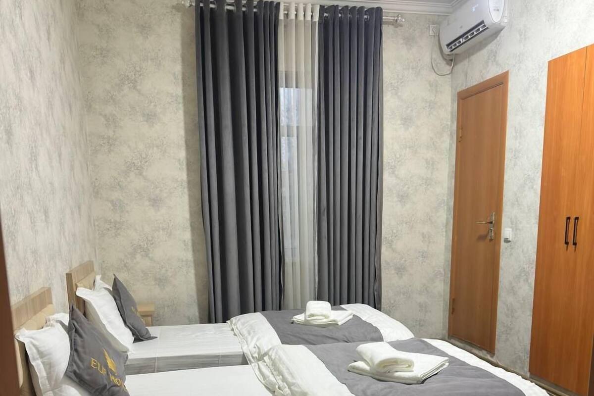 A hotel room with two beds, a wooden wardrobe, a window with curtains, and an air conditioning unit.