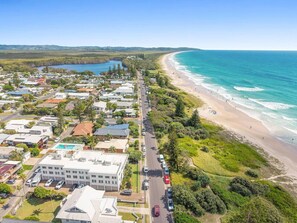 An aerial view of a coastal town with a beachfront, residential buildings, and a road running parallel to the beach. - Lennox Head Waves 2 Beachside Apartment (Lennox Head)