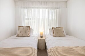 A room with two beds, a nightstand with a lamp, sheer curtains, and a view through the window. - Lennox Head Waves 2 Beachside Apartment (Lennox Head)