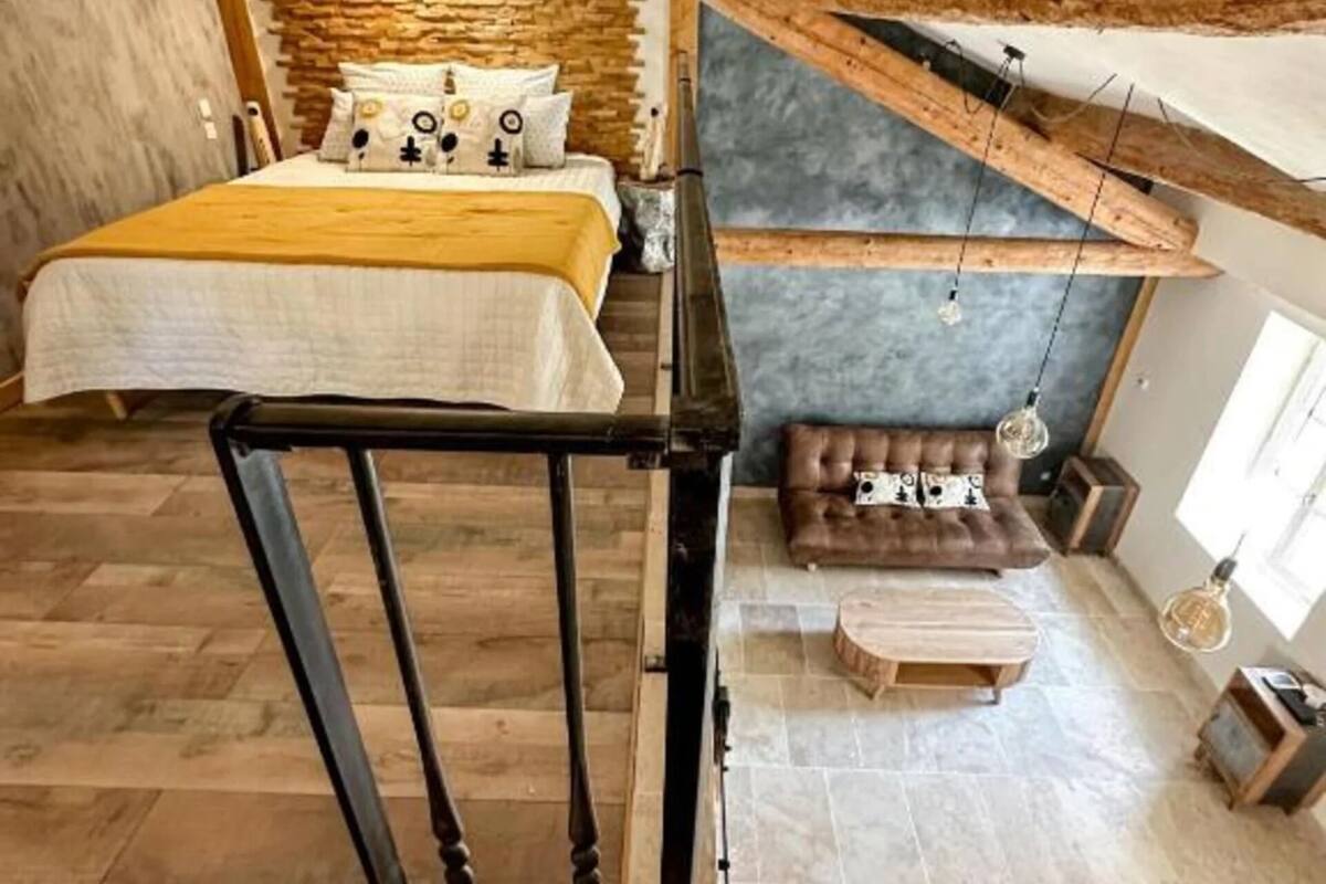 Un dormitorio tipo loft con vigas de madera expuestas, una pared de acento de piedra, una cama con colcha amarilla, un sofá de cuero marrón y una mesa de centro de madera.