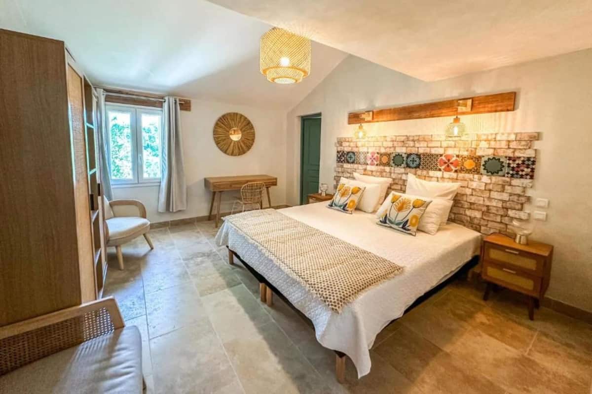 Un dormitorio con una cama grande, un escritorio y una silla. Hay una ventana con cortinas, un reloj de pared y un panel decorativo.