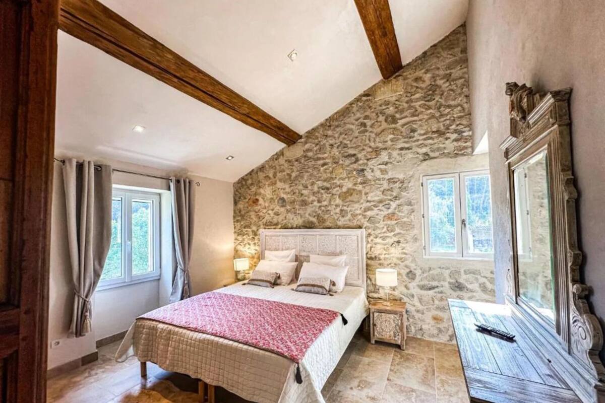 Un dormitorio con pared de piedra, techo de madera, una cama con manta roja y dos mesitas de noche con lámparas.