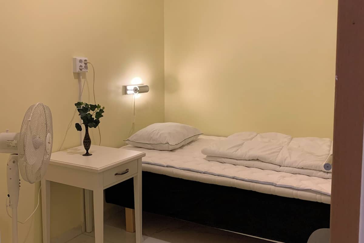 A small, well-lit room with a single bed, a bedside table with a fan, a wall-mounted light, and a power outlet.