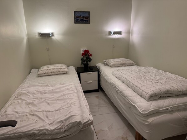 A small, clean room with two single beds, a nightstand, a small table with a vase of flowers, and a wall-mounted picture.