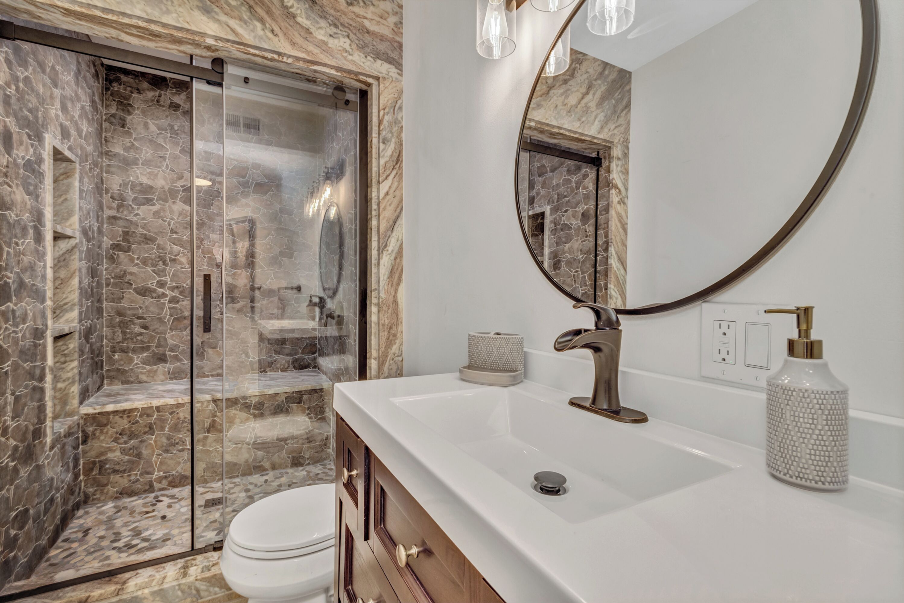 A bathroom with a stone shower enclosure, a white sink with a gold faucet, and a round mirror.