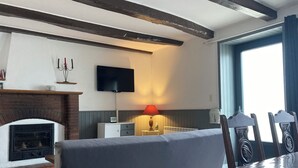 Ein gemütliches Wohnzimmer mit Kamin, einem an der Wand befestigten Flachbildfernseher, einem Esstisch mit Stühlen und einem Fenster mit Vorhängen.