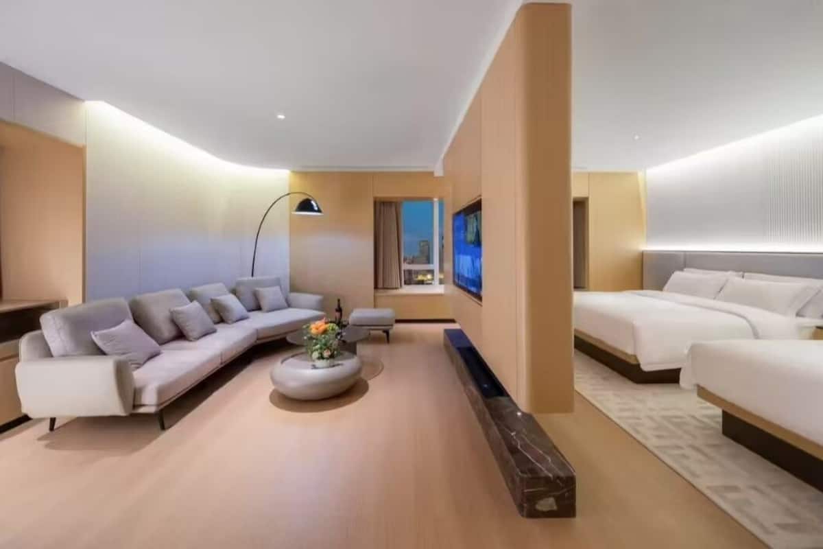 A modern hotel room with a sofa, two beds, a small table with flowers, and a large window.