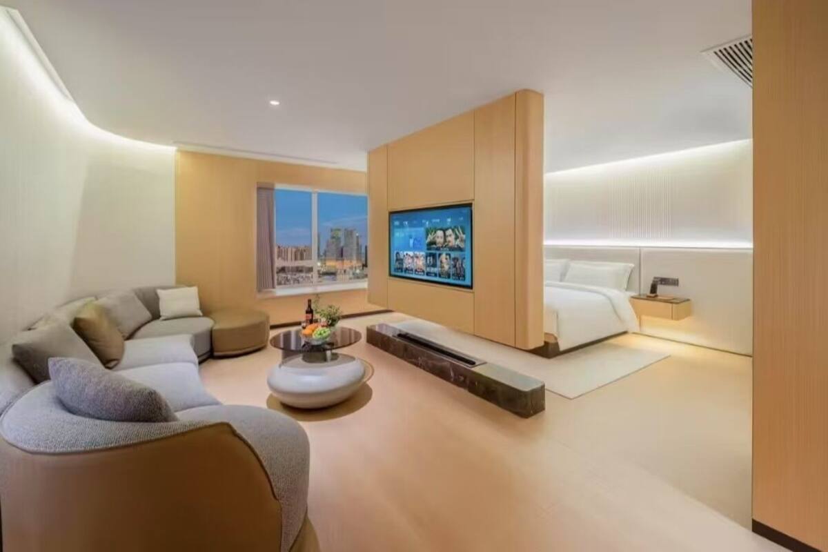 A modern hotel room with a curved wall, a large flat-screen TV, a sofa, a coffee table, and a bed.