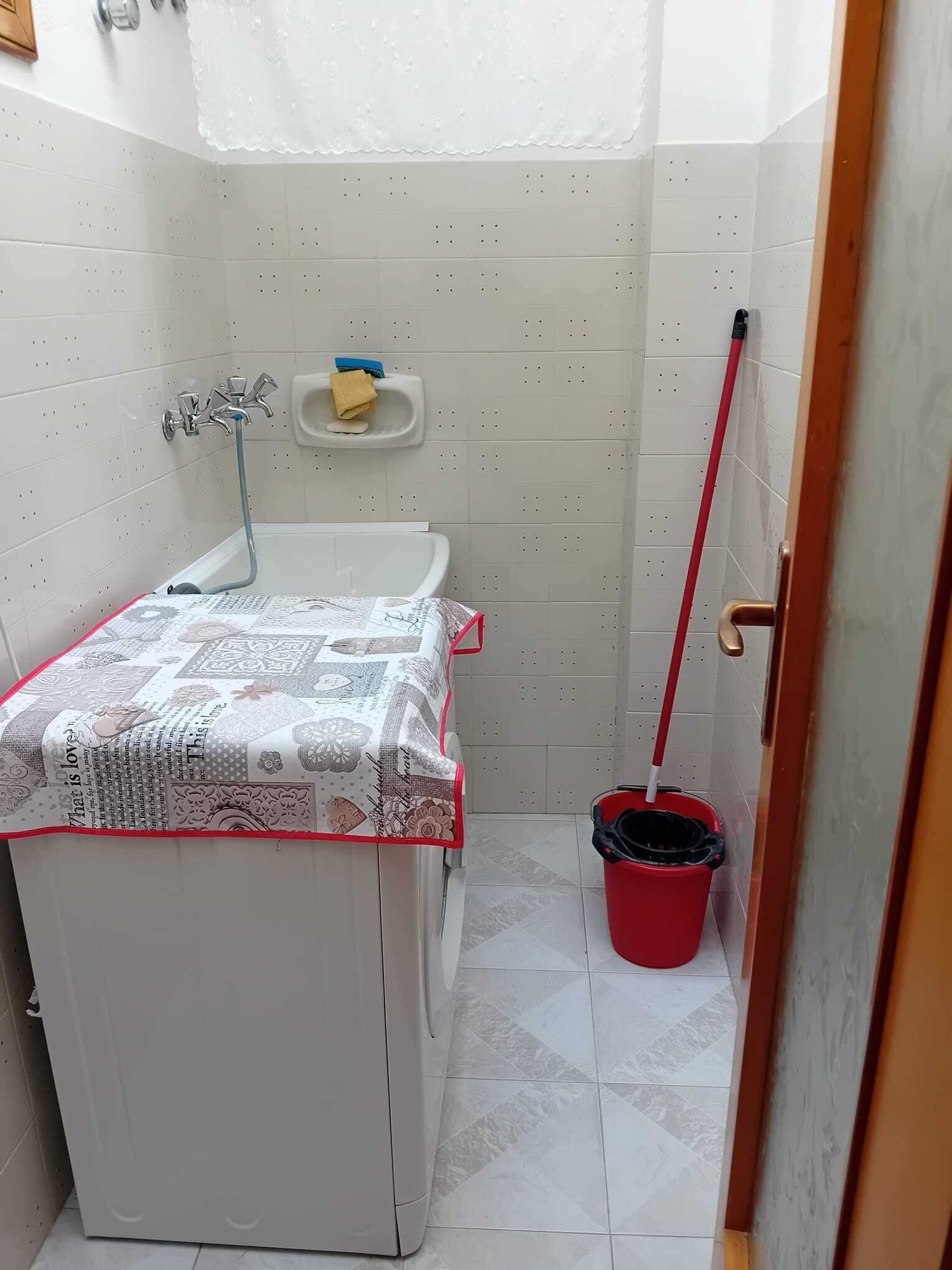 A small, clean bathroom with a toilet, a sink, and a shower. There is a red bucket and a mop leaning against the wall.