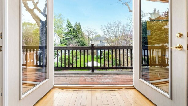 Uitzicht vanuit een schuifdeur op een houten terras met een zwarte balustrade, dat uitkomt op een achtertuin met een gazon en bomen.