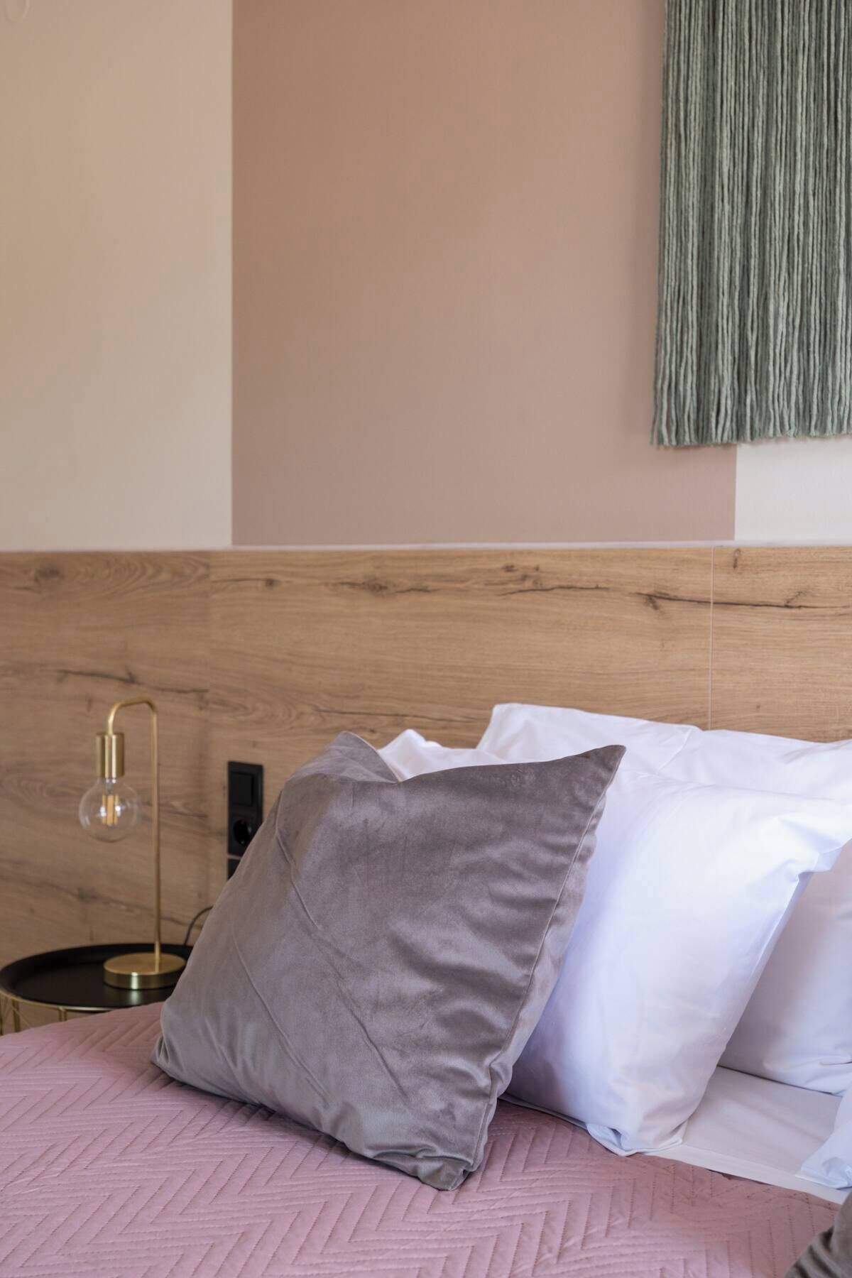 Un lit avec une couette rose, un oreiller gris et des oreillers blancs. Il y a une table de chevet avec une lampe et une prise électrique.
