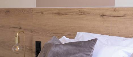 Un lit avec une couette rose, un oreiller gris et des oreillers blancs. Il y a une table de chevet avec une lampe et une prise électrique.