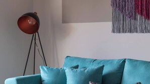 A teal sofa with multiple cushions, a floor lamp, and a wall-mounted macrame piece.