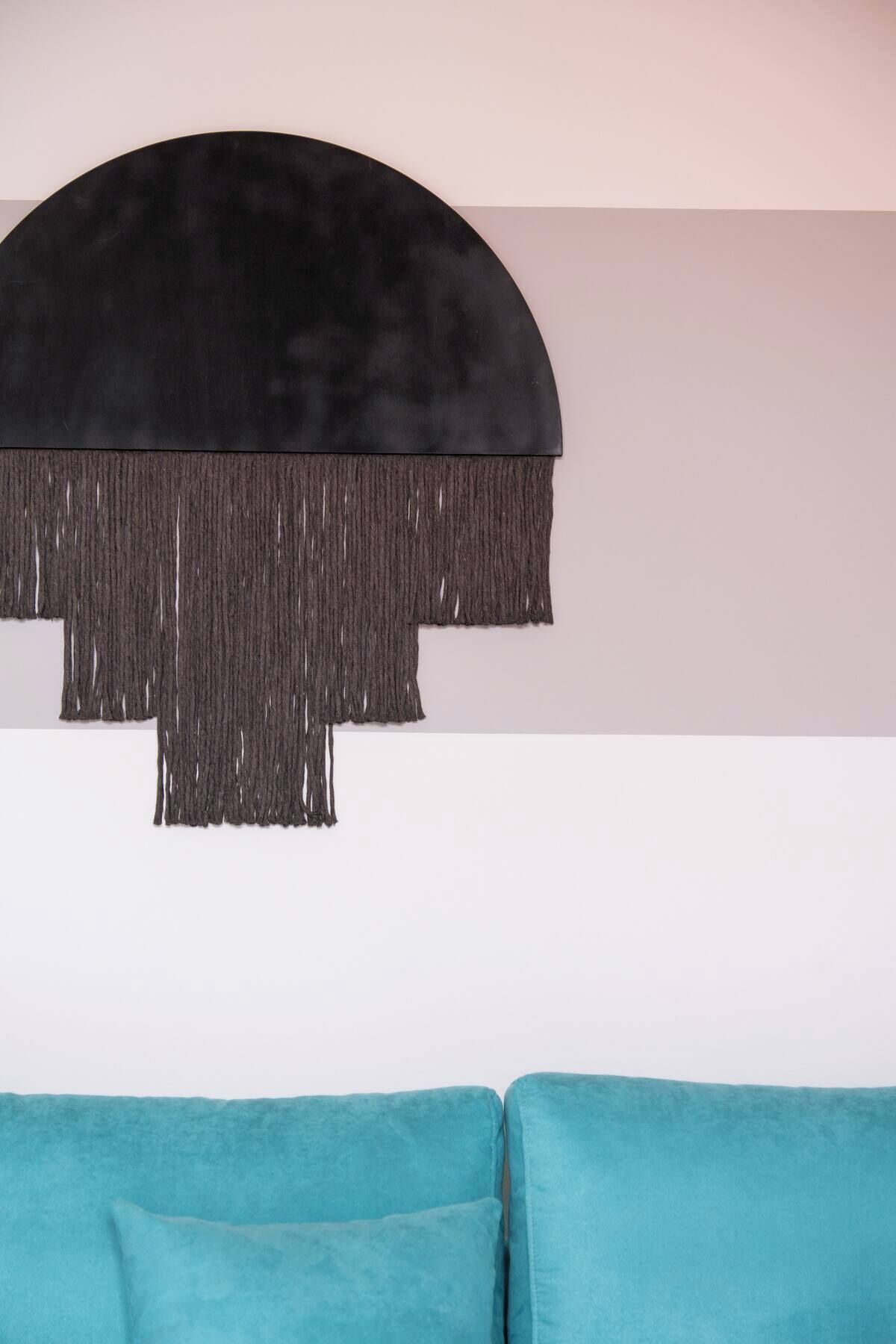 A black, semi-circular wall hanging with fringe details hanging above a teal sofa.