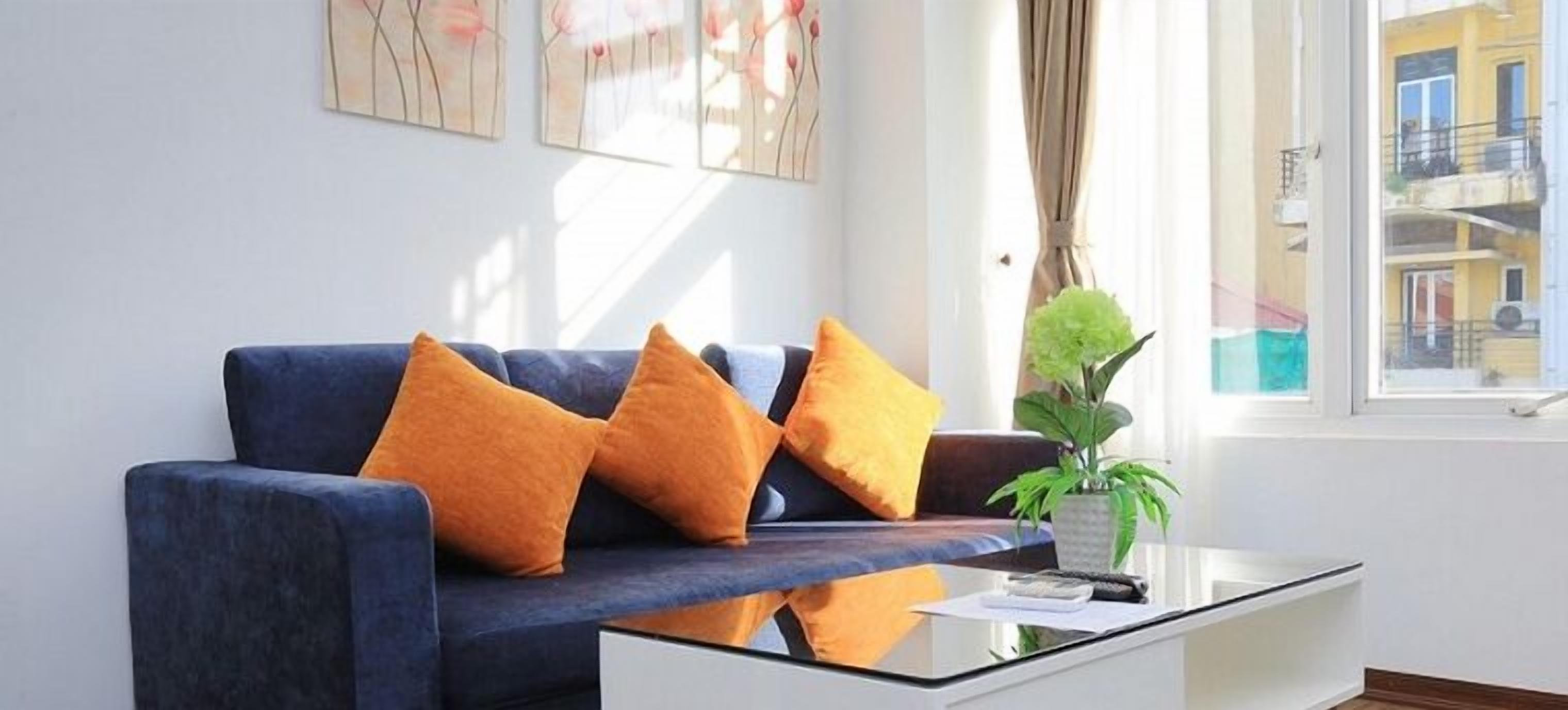 Ein modernes Wohnzimmer mit einem dunkelblauen Sofa, orangefarbenen Kissen, einem Glastisch und einer Topfpflanze.