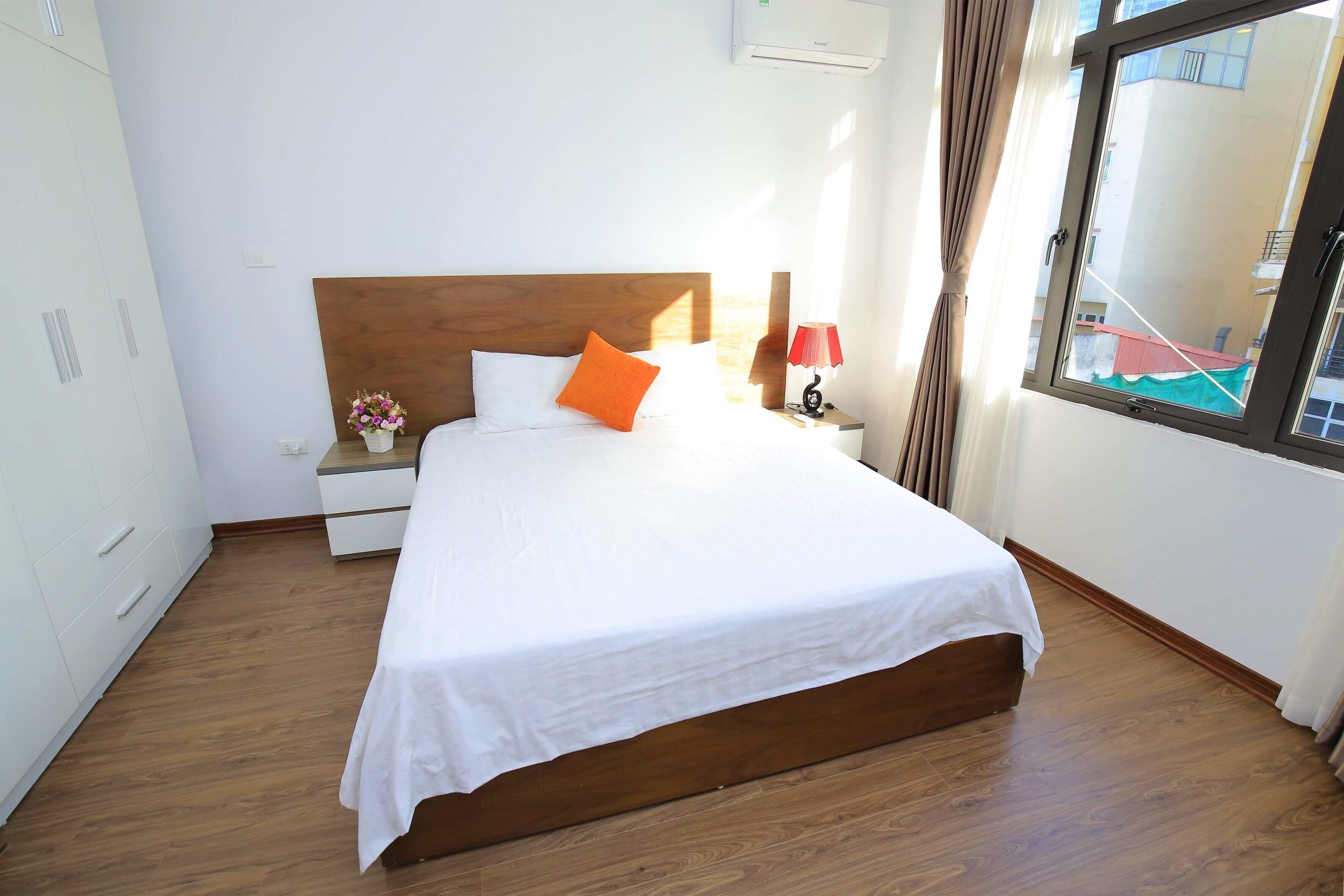 Una cama bien tendida con cabecera de madera, una mesita de noche con lámpara y una ventana con cortinas.