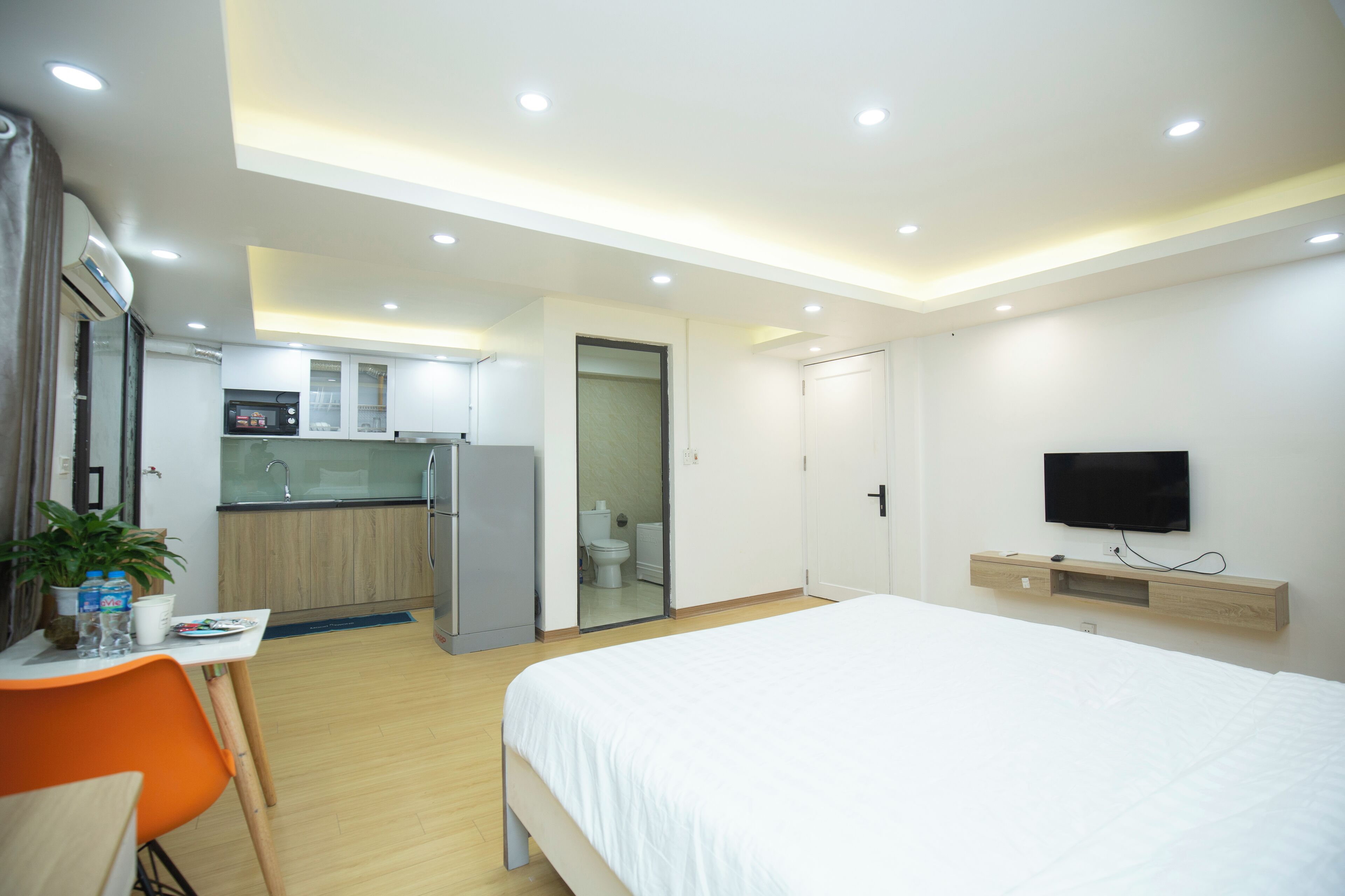 A modern hotel room with a bed, a desk, a chair, a TV, and a kitchen area.