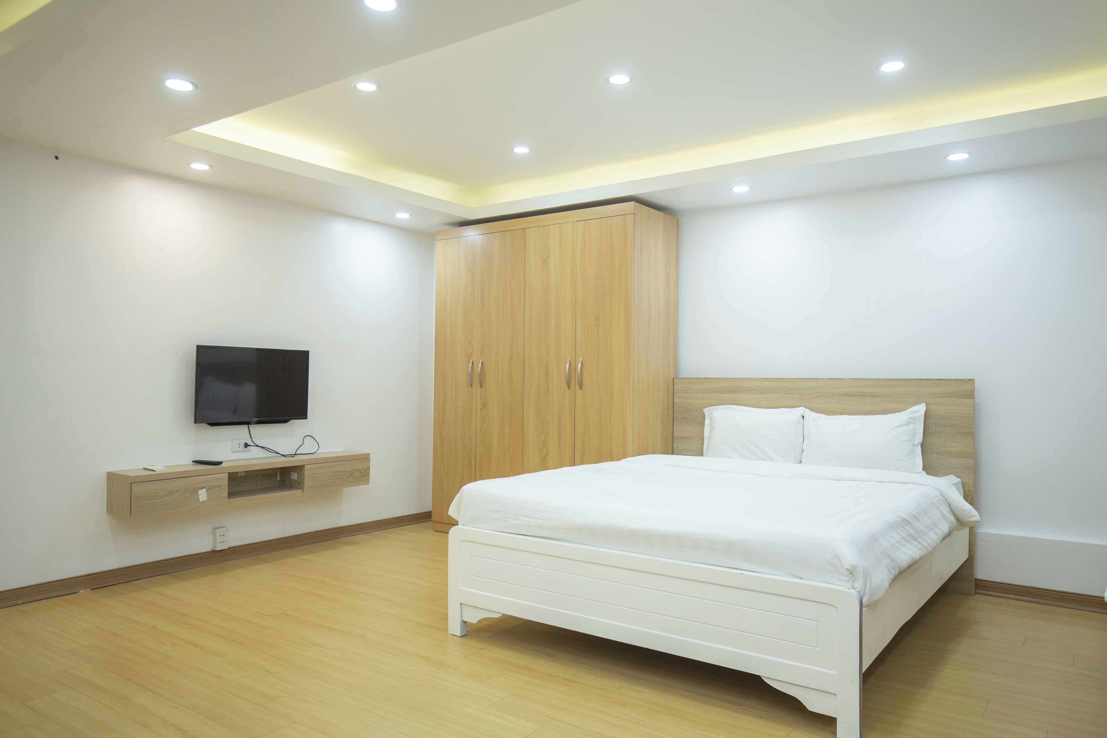 Un dormitorio moderno con cama, armario de madera, televisor integrado y luces empotradas en el techo.