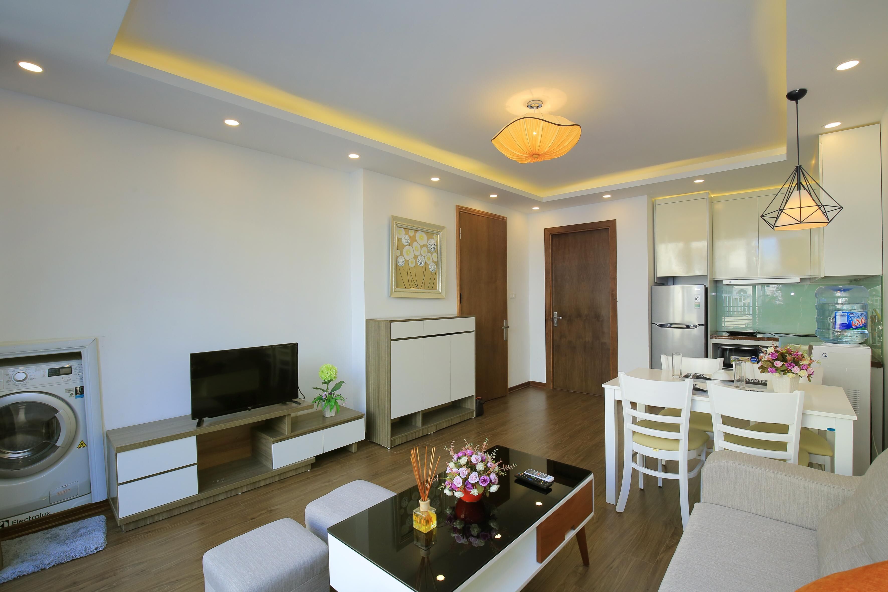 A modern living room with a flat-screen TV, a coffee table with decorative items, a dining area with a white table and chairs, and a kitchen area with a refrigerator and microwave.