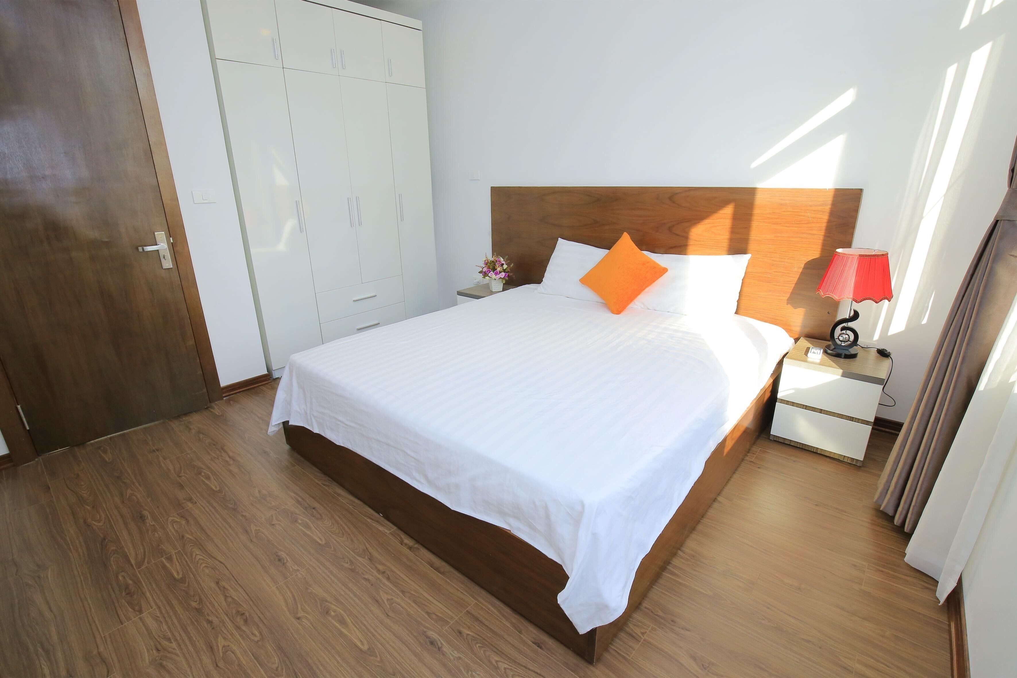 Una cama bien hecha con sábanas blancas y un almohárraga naranja, un cabecero de madera, una mesita de noche con una lámpara y un armario con espejo.