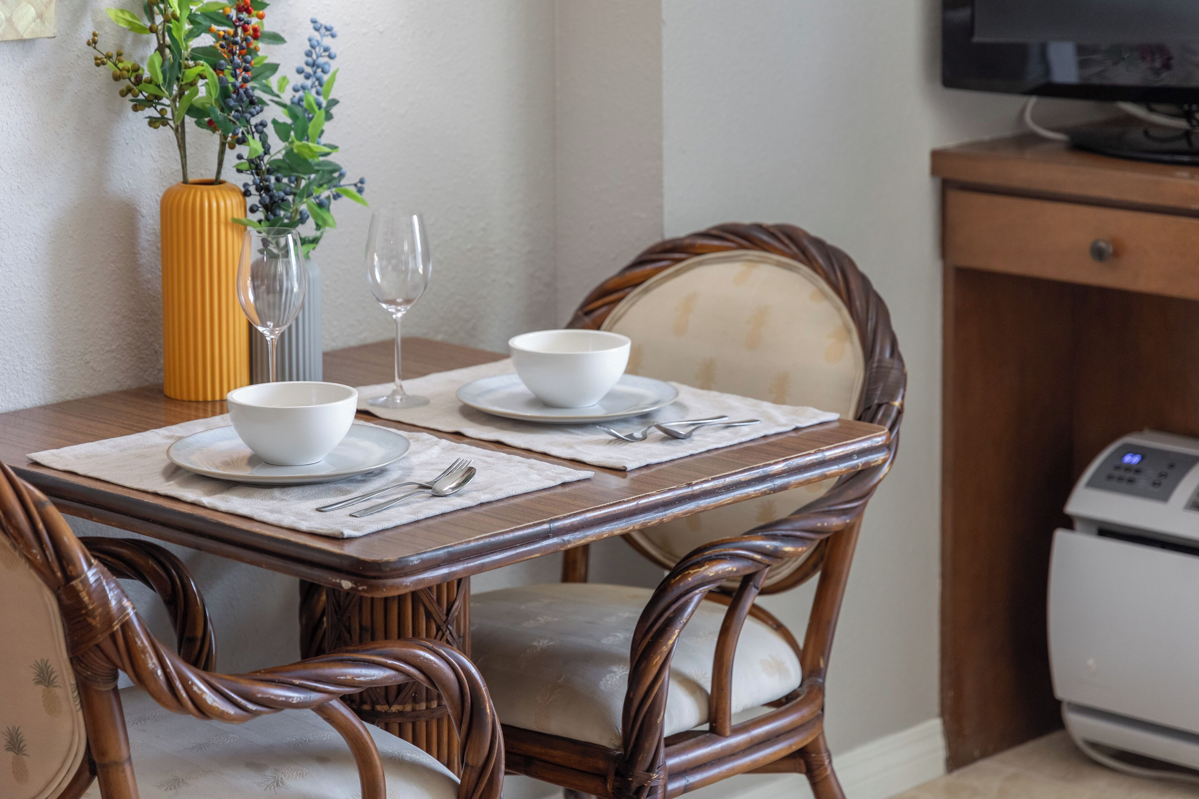 A dining table with white bowls and cutlery, a vase with flowers, and wicker chairs.