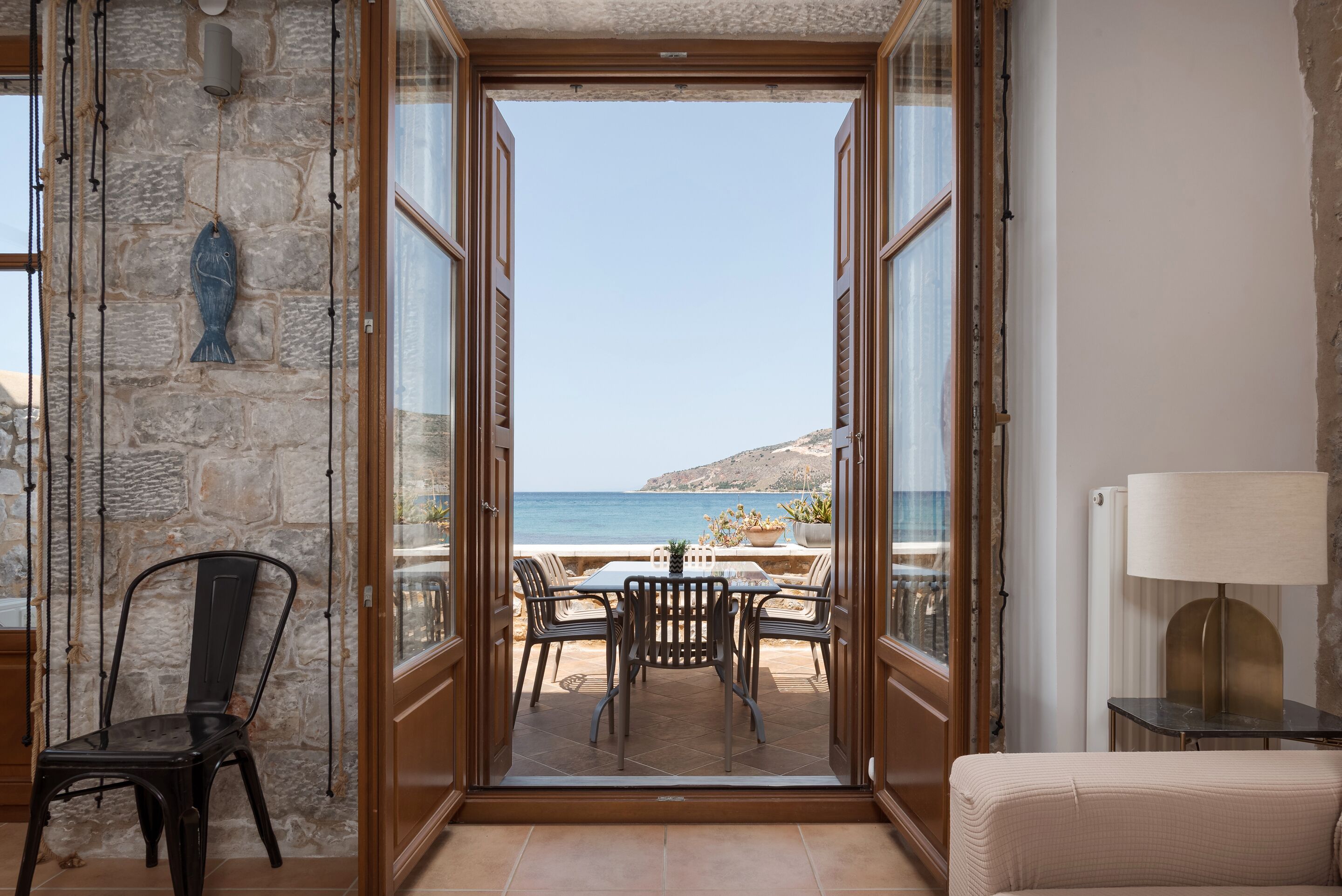 An open doorway leading to a balcony with a table and chairs, overlooking a coastal landscape.