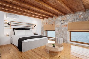 A bedroom with a bed, bedside tables, a round wicker table, a potted plant, and a stone wall with a window overlooking the sea. - Aura Seafront Villa (Neo Itilo)