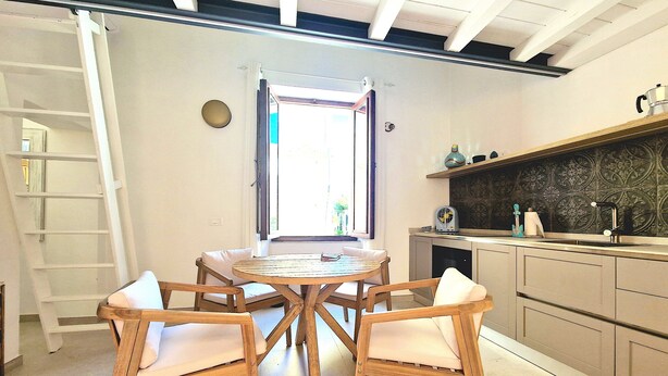 Een keuken met een ronde houten tafel en vier stoelen, een raam met uitzicht, en een inbouwplank met keukengerei.