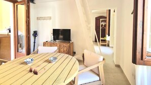 Een kamer met een houten eettafel, witte fauteuils, een televisie en een trap.
