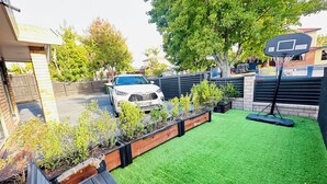 A residential backyard with artificial grass, a basketball hoop, and a white car parked in the driveway.