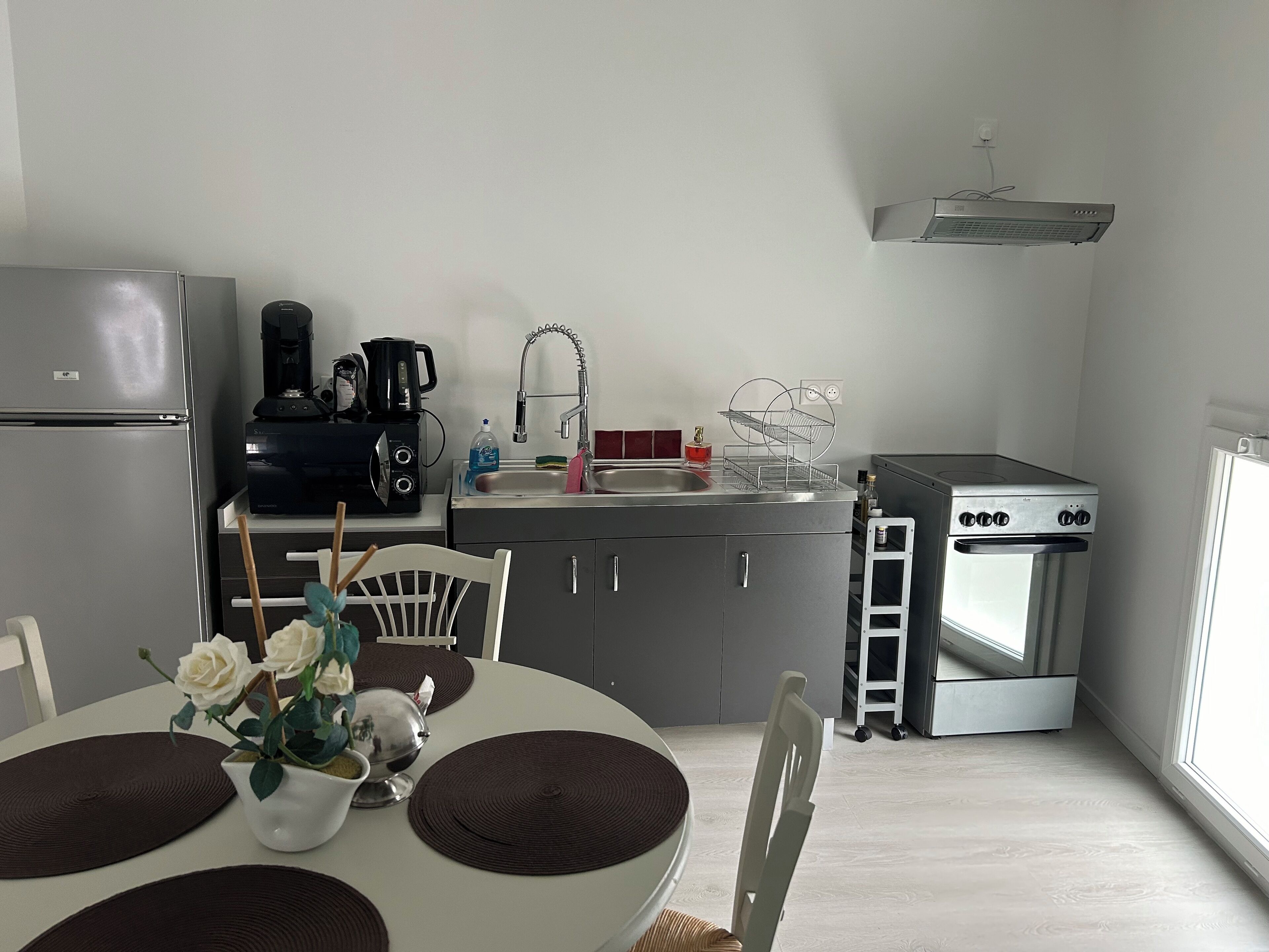 Een kleine keuken met een koelkast, koffiezetapparaat, spoelbak, fornuis en een eethoek met een tafel en stoelen.