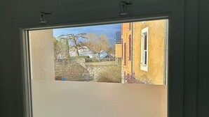 Une fenêtre donnant sur un mur de pierre, un arbre et un bâtiment aux ornements rouges.
