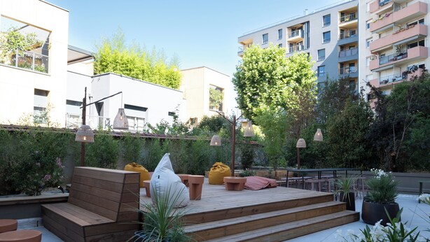 Un espace extérieur aménagé en terrasse, avec des sièges en bois, des plantes en pot et une vue sur les bâtiments environnants.
