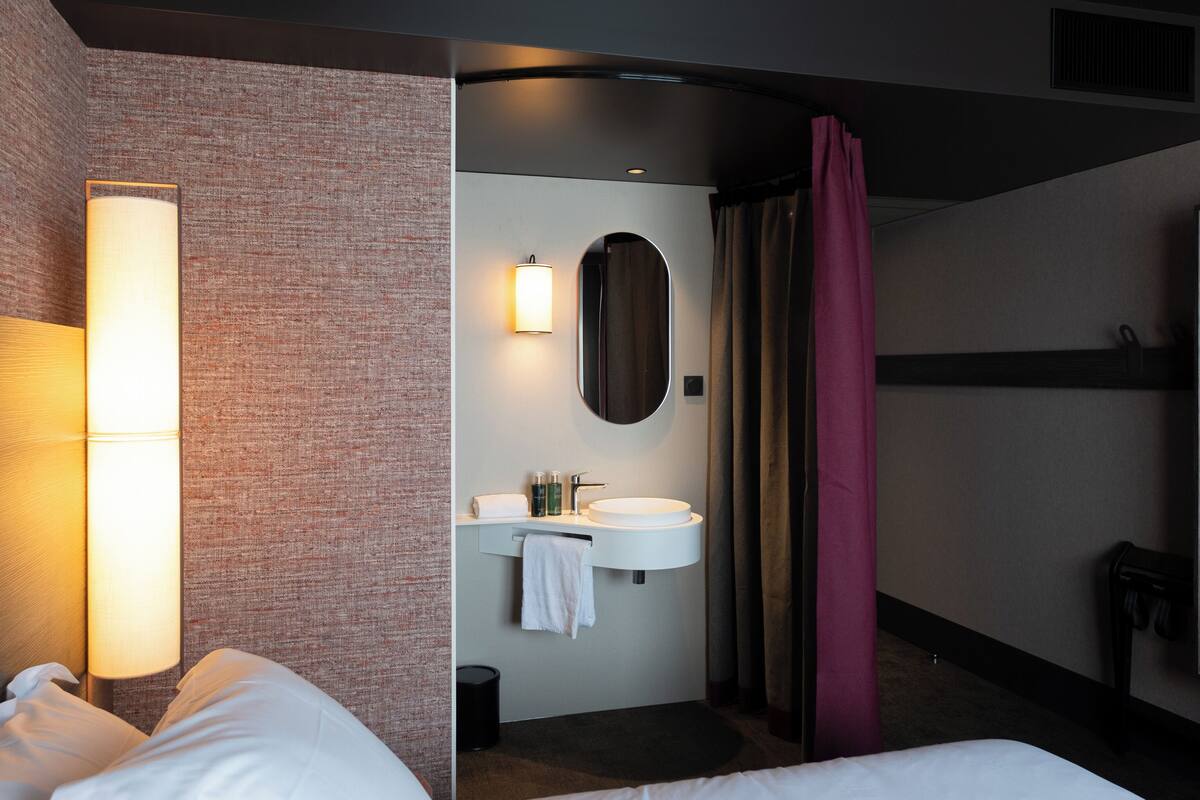 A hotel room with a bed, a bedside lamp, a bathroom with a sink, mirror, and towel rack.