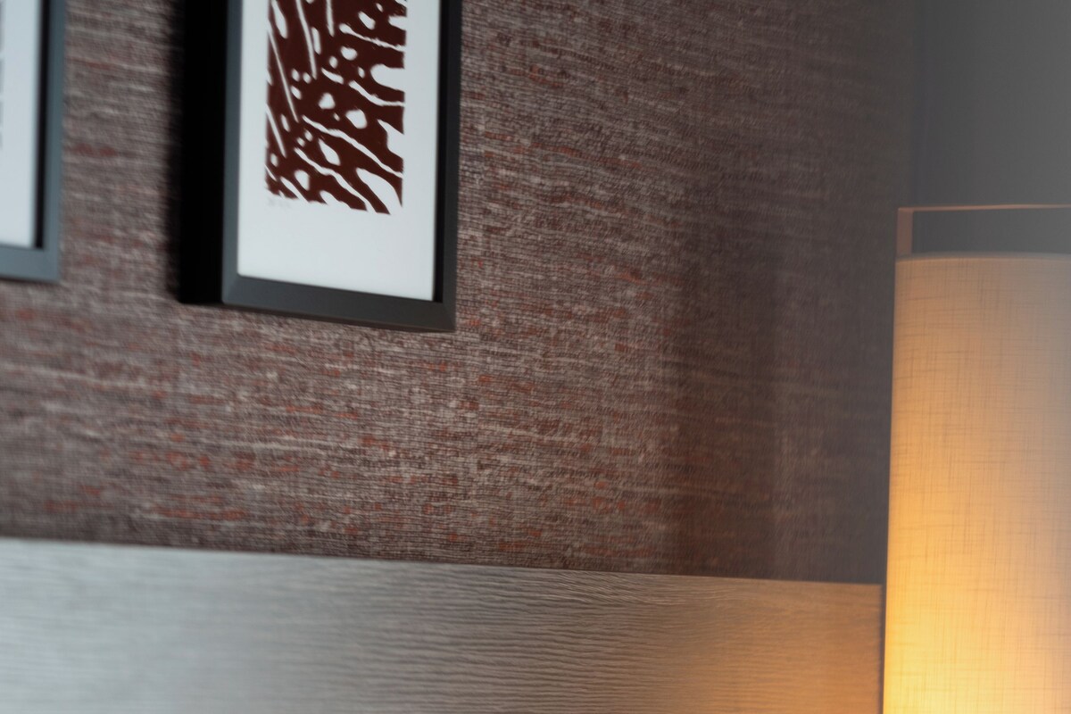 A neatly made bed with white linens, a textured wall, and a wall-mounted lamp.