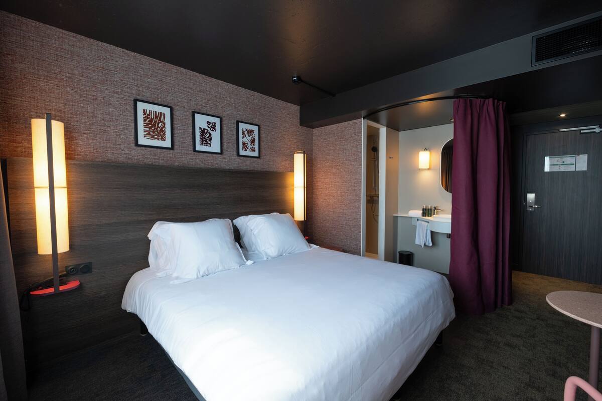 A hotel room with a large bed, two wall-mounted lamps, a mirror, a small table, and a bathroom visible through an open door.