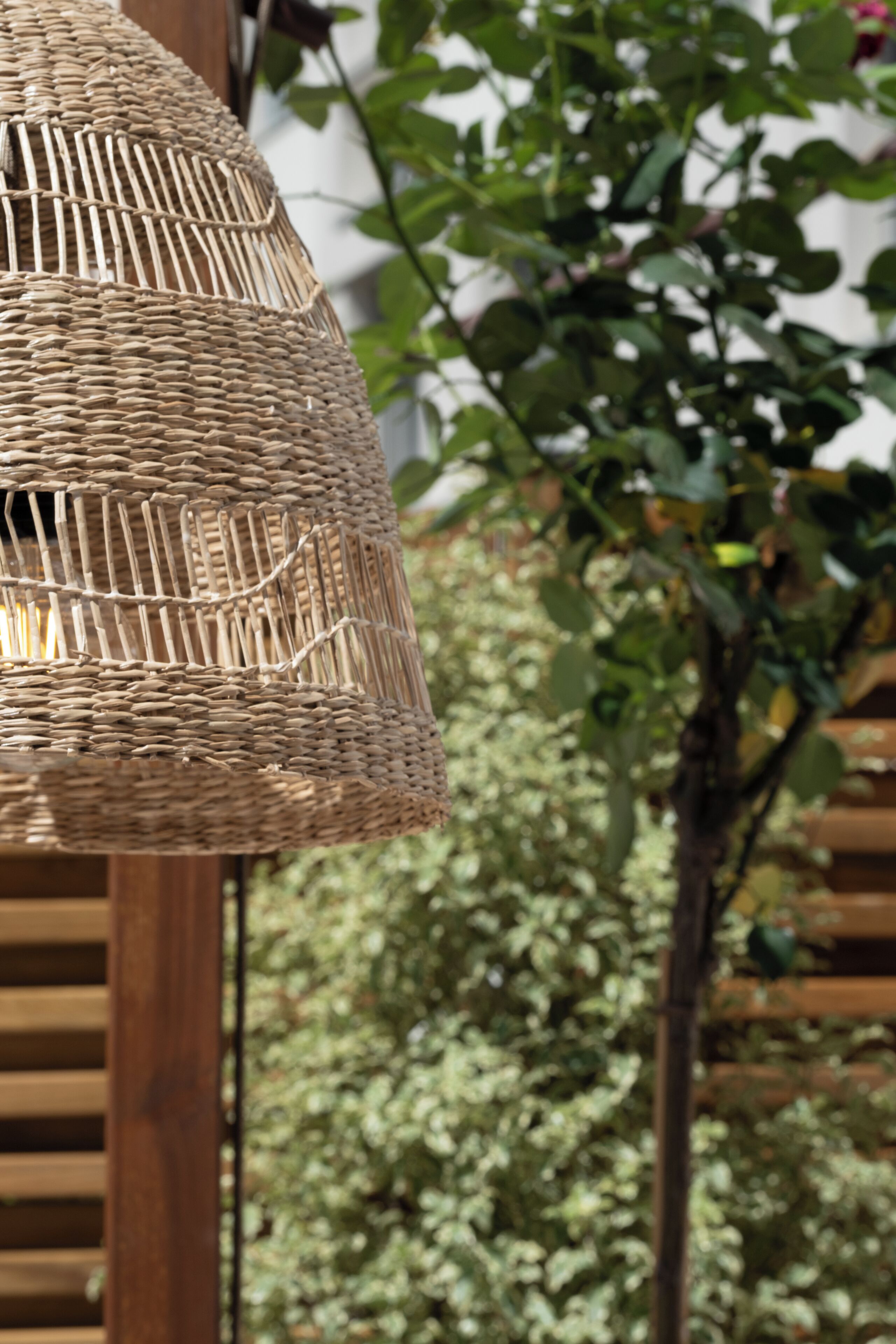 Una lámpara de mimbre con base de madera, frente a un fondo de follaje y una estructura de madera.